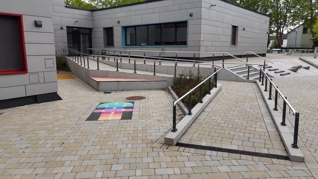 Eine Rampe mit Gel&auml;nder und ein bunt aufgemaltes H&uuml;pfspiel auf dem Pflaster zwischen zwei Geb&auml;udeteilen
