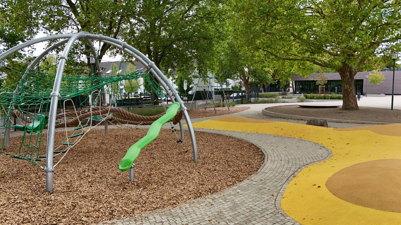 Ein Kletterger&uuml;st aus Stahlb&ouml;gen mit gr&uuml;nem Netz und gr&uuml;ner Rutsche, darunter Rindenmulch, daneben eine gelbe Spielfl&auml;che