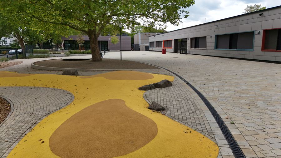 Der Schulhof mit einer runden Sitzstufe um einen gro&szlig;en Baum, gelben Bodenfl&auml;chen und dem langen Geb&auml;uderiegel dahinter
